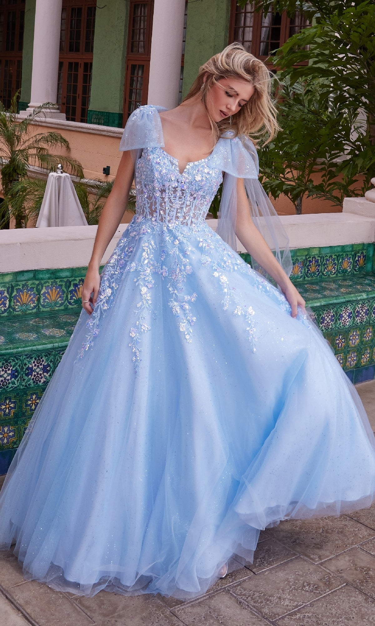 A woman in the Nox Anabel H1680 Shoulder-Bows Long Prom Dress, featuring a light blue floor-length skirt and sheer cape sleeves, stands outdoors by a green tiled wall.